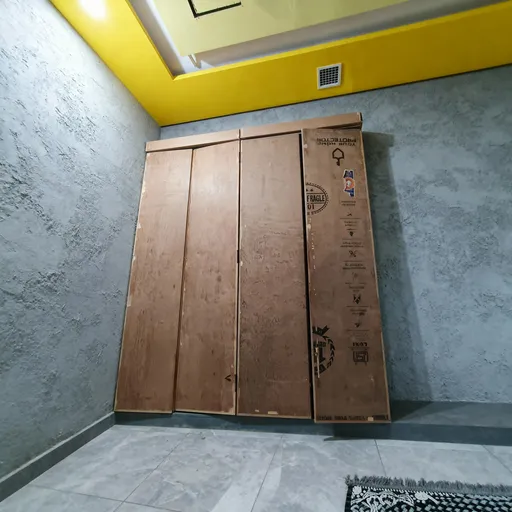 Prompt: The photograph depicts a set of large, brown cardboard storage boxes stacked against a wall in what appears to be a basement or storage room. The boxes are tall and rectangular, with visible black text and symbols printed on their surfaces, including "FRAGILE" and various other markings. The box on the far right has a visible handle on the side and a few small stickers, including a triangular warning sign. The boxes are positioned vertically, with the edges aligned and the tops aligned horizontally. The wall behind the boxes is painted gray and has a slightly rough texture. Above the boxes, the ceiling is painted bright yellow, contrasting with the gray wall and brown boxes. A small, rectangular air vent is visible in the ceiling. The floor is covered with gray tiles, and a black and gray braided rug is partially visible in the bottom right corner of the image. The lighting is bright and even, suggesting that the room is well-lit. The overall scene conveys a sense of organization and storage, with the boxes serving as a means to keep items protected and organized in a functional space.