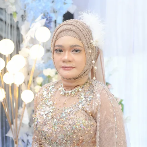 Prompt: This photograph captures a young Southeast Asian woman standing indoors against a backdrop of soft, white curtains and an arrangement of white and blue flowers. She is dressed in a traditional, elaborate bridal outfit, known as a kebaya. The kebaya is a sheer, beige garment adorned with intricate, shimmering sequins and beads that catch the light, creating a dazzling effect. The bodice is fitted and the sleeves are three-quarter length, both covered in the same sparkling embellishments. Her head is covered with a beige, netted hijab that features a white, feathery adornment on the right side, adding a touch of elegance. She wears a matching choker necklace with similar sequins and beads. Her makeup is subtle, with a focus on her eyes, which are accentuated with eyeliner and eyeshadow. The background includes a set of tall, round, white light bulbs on the left, adding a soft, ambient glow. The overall atmosphere is one of elegance and festivity, with a focus on traditional bridal beauty and celebration. The photograph is well-lit, highlighting the intricate details of her outfit and the soft, decorative elements around her.
