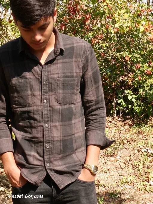 Prompt: Dressed in a dark plaid shirt, a young man stands outdoors, his head bowed slightly as he appears introspective or contemplative. His hands rest in the pockets of his black jeans, while one wrist glimmers with a gold watch, adding a touch of elegance to his casual attire. The backdrop reveals a lush, green environment, suggesting a warm day illuminated by bright sunlight, enhancing the vivid colors of the foliage around him. The composition focuses on his thoughtful posture and relaxed demeanor, creating a mood of tranquility and reflection in this serene outdoor setting.