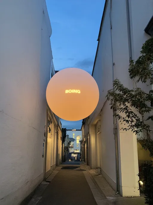 Prompt: A massive, glowing orb hangs precariously between two narrow white buildings in an urban alley, casting a warm, amber light that diffuses softly against the surrounding surfaces. Written on the orb are the words "BOING", is fat cartoonish letters.  The sky above transitions to a deeper blue as dusk settles, providing a striking contrast to the vibrant scene below. Strands of decorative lights dangle from each side of the alley, illuminating the wet pavement which reflects the lights, adding a touch of intimacy to the space. The whimsical installation of the orb, while the distant glow of other buildings hints at a lively atmosphere.