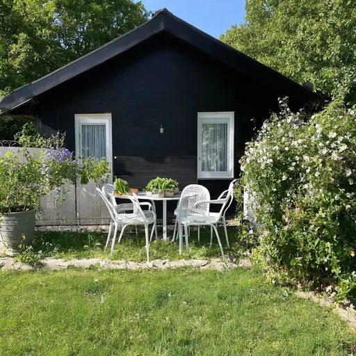 Prompt: Garten mit einem staketen Zaun und vielen Blumen im Hintergrund ein kleines Holzhaus. Das Haus hat ein schwarzes Dach und einen schwarzen Anstrich mit weißen Fenstern und einer weißen Tür. Vor dem Holzhaus ist eine Terrasse mit einem weißen Tisch und vier weißen Stühlen. Um die Terrasse sind viele Blumen . Es liegt Gemüse in einem Weidenkorb auf dem rasen hinter dem Zaun 