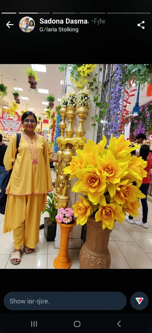 Prompt: In a vibrant marketplace filled with colorful decorations, a woman stands confidently next to an elaborate display of artificial flowers and plants. Dressed in a flowing yellow kurta with delicate pink embellishments, she exudes a warm presence, her glasses framing a gentle smile. The backdrop features a plethora of floral arrangements and ornate gold items, creating an inviting atmosphere rich with festive energy. Bright yellow flowers dominate the front, complemented by a variety of colorful blooms and greenery, enhancing the scene's cheerful aesthetic. The overall ambiance is lively and engaging, suggesting a cheerful shopping experience amid the celebrations.