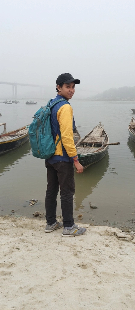 Prompt: Standing by the water's edge, a young man with a playful smile faces the camera, dressed in a colorful jacket with blue and gold accents, complemented by a black cap. He carries a large teal backpack, which adds a vibrant touch against the muted tones of the surrounding misty atmosphere. Behind him, wooden boats gently bob in the calm waters, partially obscured by fog, and a distant bridge looms on the horizon, hinting at an expansive view. The sandy shore beneath his feet is scattered with occasional debris, enhancing the natural, rugged ambiance of the riverbank scene. The overall mood is tranquil yet lively, capturing a moment of casual exploration along the water's edge.
