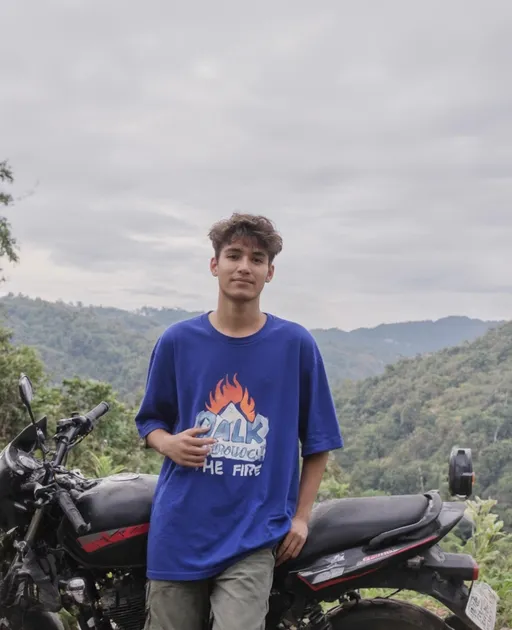 Prompt: Standing confidently in a lush, hilly landscape, a young man in a vibrant blue oversized t-shirt emblazoned with "WALK THROUGH THE FIRE" strikes a casual pose beside a black motorcycle with red accents. His tousled hair and calm expression suggest a relaxed demeanor, harmonizing with the tranquil surroundings, which are bathed in soft, diffused light under a cloudy sky. The background features a mix of greenery and distant hills, creating a serene, almost dreamlike ambiance, while the motorbike adds an adventurous touch to the scene. This image captures a moment of youthful exploration, underscored by the simplicity of nature and the quiet strength of the subject.
