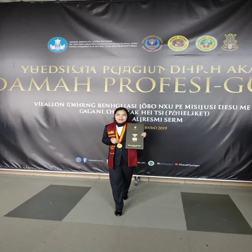 Prompt: This is a photograph of a woman standing in front of a large black banner with gold and white text in Indonesian. The banner reads "YUDISIUM PENGKUKUHAN DAUMAH PROFESESIG" and lists the names and affiliations of participants and organizers. The woman is centered in the image, standing on a gray-tiled floor with two dark gray rectangular tiles positioned at her feet. She is wearing a formal black suit with a red and gold sash draped over her left shoulder, and a maroon tie. She has a gold medal hanging from a red ribbon around her neck, and she is holding a silver plaque in her right hand. Her hair is neatly tied back, and she is wearing black shoes. The background features multiple logos and emblems at the top of the banner, including a blue circular logo with a white bird figure and a yellow circular emblem with a red and blue design. The overall setting appears to be indoors, likely in a conference or event hall, given the formal attire and the structured banner display. The lighting is bright, highlighting the woman and the banner text clearly.