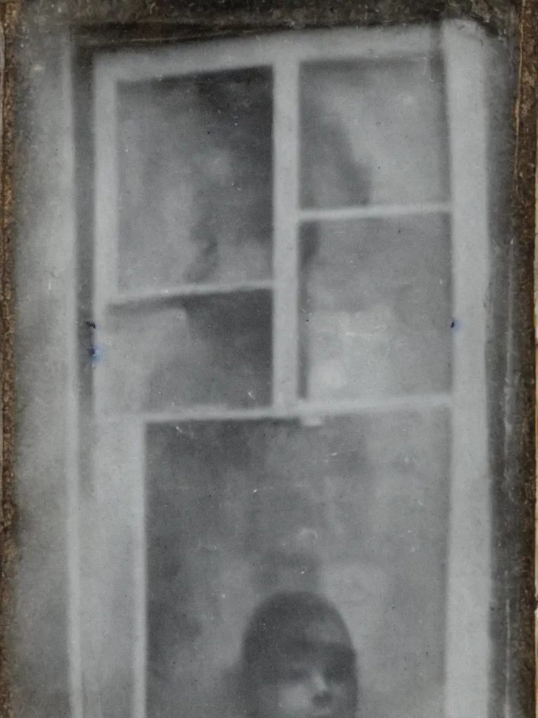 Image similar to a tintype photograph. the face of a ghost in the window of a house, next to the front door