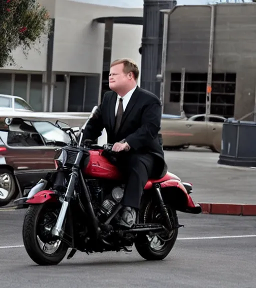 Image similar to Andy Richter is dressed in a black suit and a red necktie and riding a motorcycle into a studio lot. The studio lot is filled with Soundstages and movie trailers. It is a bright afternoon and overcast.