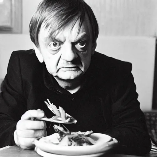 Prompt: mark e smith sitting at a table eating a big pile of vegetables, fork and knife in hand, drooling and licking and smacking his lips, photograph