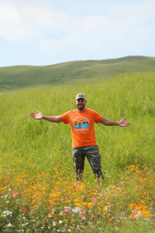 Prompt: Amidst a vibrant landscape of lush green grass and colorful wildflowers, a man stands with arms outstretched in a gesture of joy and freedom. Dressed in a bright orange T-shirt featuring a scenic logo, camouflage pants, and a cap, he exudes a sense of adventure and contentment. The backdrop showcases rolling hills under a sky that hints at a gentle overcast, creating a tranquil yet invigorating atmosphere. Patches of pink and yellow flowers bloom at his feet, adding splashes of color that enhance the feeling of being immersed in nature. The overall scene captures a spirit of exploration, celebrating the beauty of the outdoors and the exhilaration of being in a wide-open space.